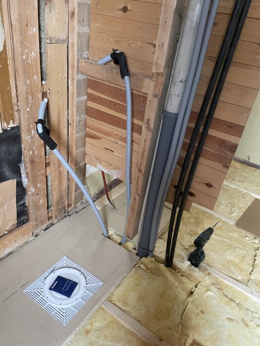 Byggprojekt med vägg under renovering, nya rörinstallationer och isolering synliga. Borra och verktygslåda på golvet vid arbetet.