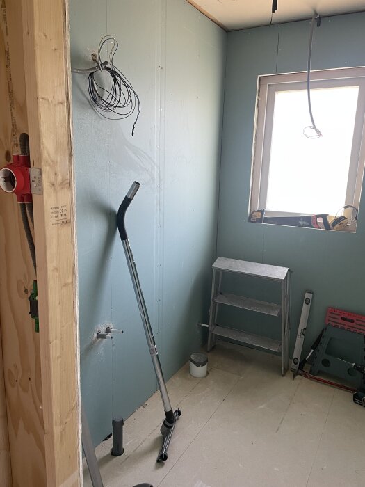Ett pågående renoveringsprojekt med nya väggar, stege, verktyg och synliga rör i ett ljust rum med fönster.