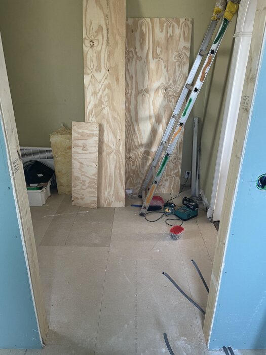 Rum under renovering med stege, plywoodskivor och verktyg på golvet, vita väggar och blå dörrkarm. Elektriska kablar syns på golvet.
