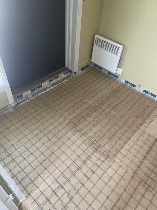 Golv med nät av metalltrådar och en väggmonterad vit värmepanel i ett rum under renovering.