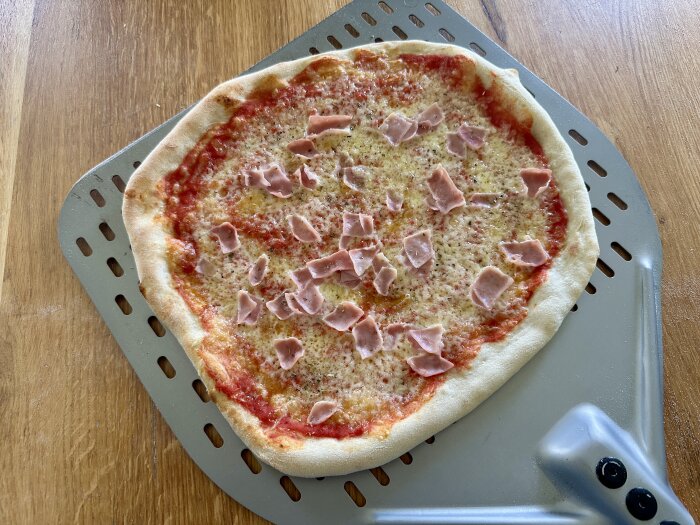 Hemlagad pizza med tomatsås, ost och skinka, upplagd på en pizzaspade av metall på ett träbord.