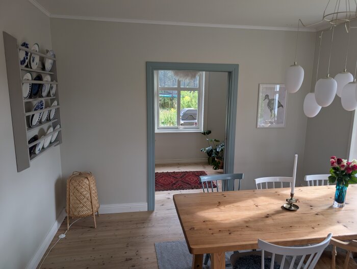 Matrum med träbord och stolar, en tallrikshylla på väggen, och en öppning till ett rum med fönster, krukväxt och röda mattor.