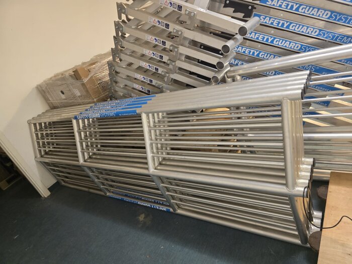 SGS fallskyddspaket för lutande tak, 24 meter, nytt och oanvänt. Staplade metallramar och komponenter i ett rum, märkta "Safety Guard Systems".
