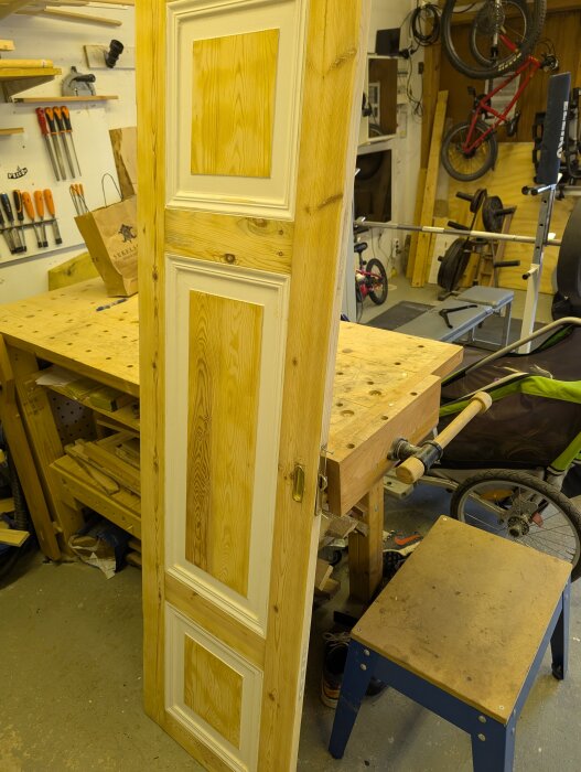 En trädörr med vitmålade paneler står lutad mot en arbetsbänk i en verkstad med verktyg och cyklar i bakgrunden.