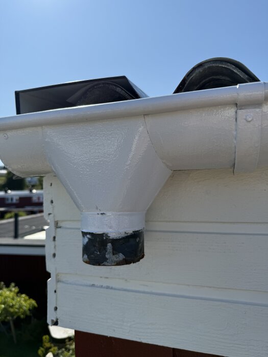 Nedre delen av ett stuprör monterat vid takets hängränna på ett hus, målat vitt, med en metallanslutning som ser ut att sitta fast.