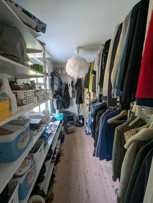 Garderob med kläder hängande till höger och hyllor med lådor och föremål till vänster. Träparkettgolv och en dekorativ taklampa syns.