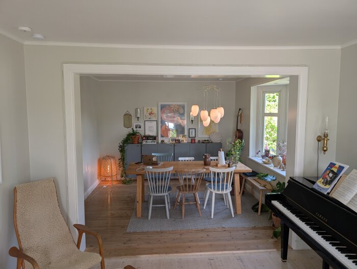 Öppet och ljust rum med träbord och stolar, piano till höger, och gröna växter vid fönstret. Väggen mellan rummen har tagits ned.