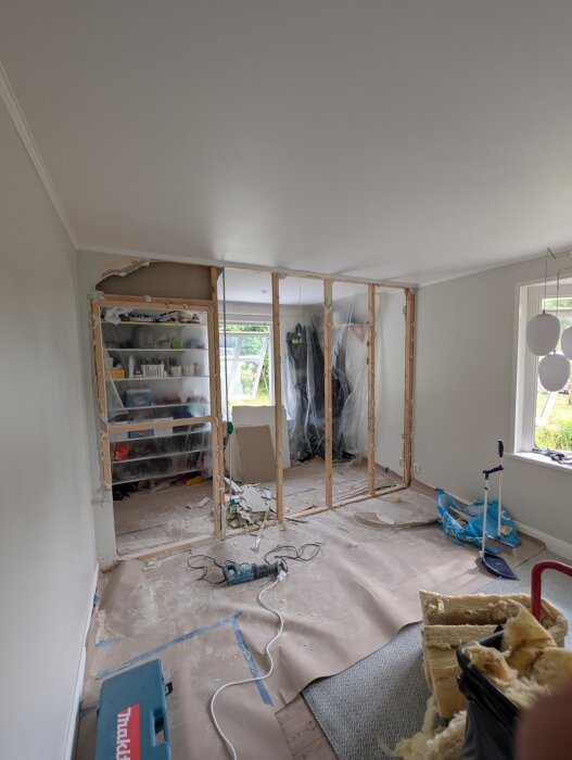 Riven innervägg med synliga reglar, byggmaterial och byggverktyg på golvet. Garderobinnehåll synligt i bakgrunden.