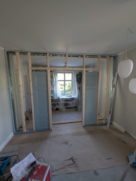 Träramar och blå skjutdörrar monterade i en nyinnervägg under byggnation, med verktyg och papper på golvet.