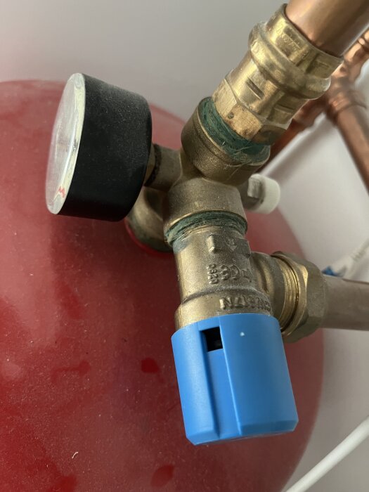 Expansionskärl i ett värmesystem med tryckmätare och blå ventil, kopplad till kopparrör, på röd bakgrund.