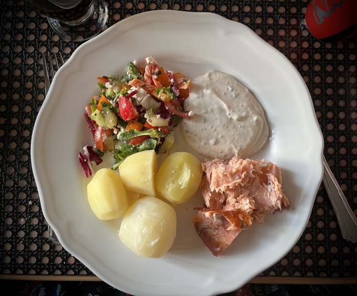 Tallrik med rökt lax, kokt potatis, sallad och romsås på ett bord.