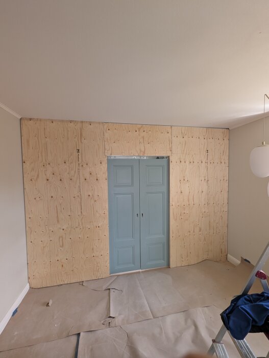 Innervägg byggd med plywood och blå skjutdörrar synliga i mitten, omgiven av monterad pärlspont och skyddspapper på golvet.