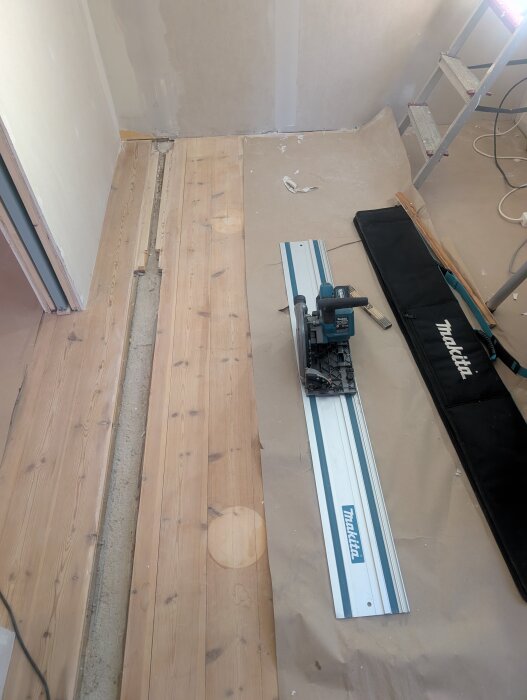Golvbjälklage inomhus under renovering, en sänksåg ligger på en träplanka bredvid ojämna golvbrädor och påbörjad golvreparation.