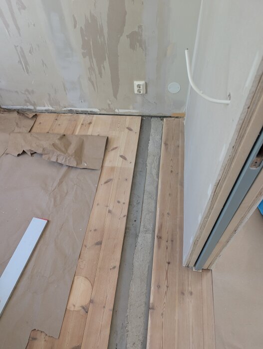Renovering av golv med delvis utbytt golvplanka, där en innervägg tidigare stod, med papper och verktyg synliga runt omkring.