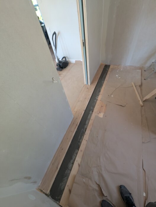 Golvlagningsprojektet med en gammal golvplanka, omgivande hål fixat med spackel och papper på plats för att skydda ytan, dammsugare i bakgrunden.