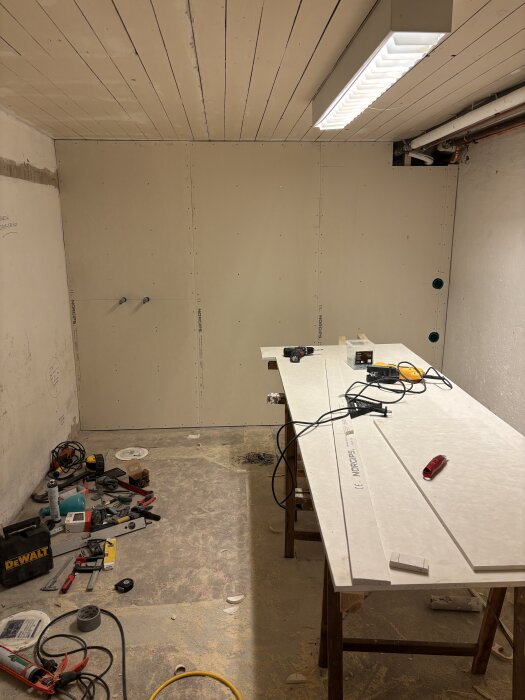 Byggarbetsplats med verktyg och material, vägg med färdig plyfa och gips, arbetsbänk med kabel och verktyg inför flytspackling och målningsarbete.