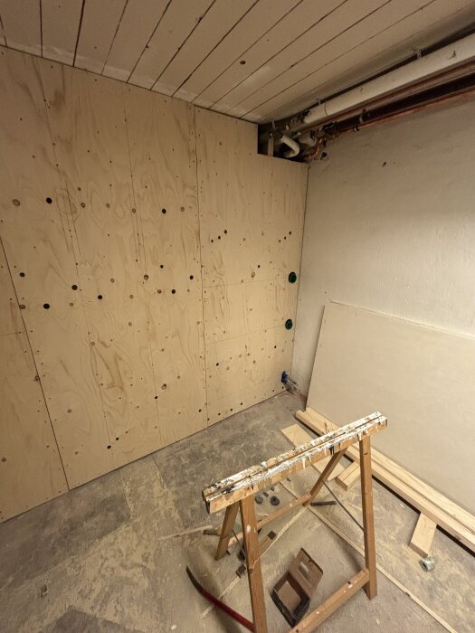 Byggprojekt med vägg av träpanel och gips redo för flytspackling. Verktyg och material på golvet; rördelar vid taket synliga.