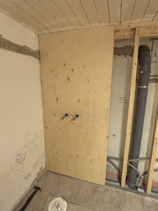 Vägg med plywoodskiva och gipsskiva, synliga rör och eluttag i ett rum under renovering. Verktyg och burk på golvet.