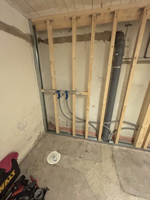 Träram för väggkonstruktion med rördragning och märkningar på betonggolv i färd med renovering. Verktyg synliga i nedre hörnet.