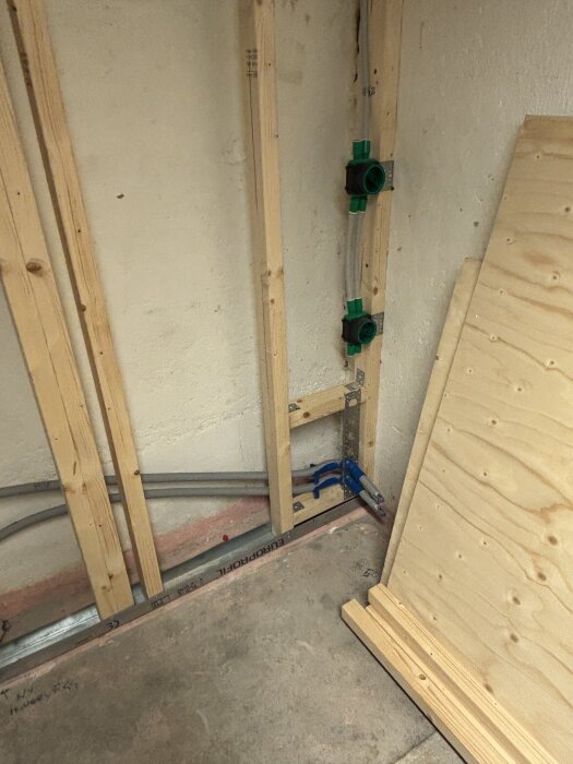 Byggarbetsplats med träreglar och synliga rör mot en vägg, plywoodskivor står lutade mot väggen.