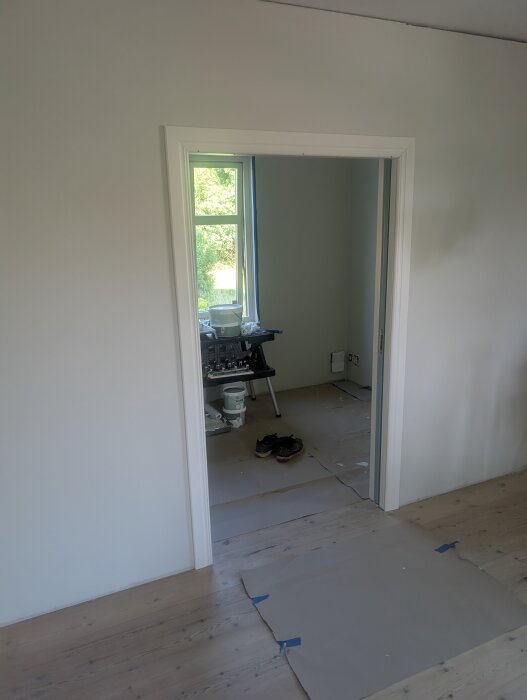 Öppning med dörrfoder, rum under renovering; golv täckt av skyddspapper, målarburkar och skor i bakgrunden, soligt fönster.