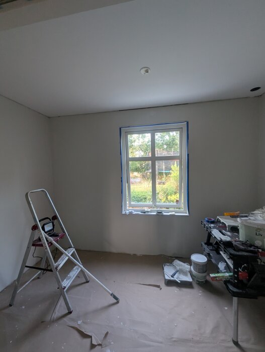 Rum under renovering med målad vägg, fönster skyddat med tejp, stege, och verktygsbord fullt av målarutrustning.