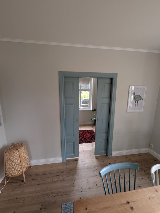 Öppna dubbeldörrar i ljusgrön nyans med en röd matta i det anslutande rummet. Trämöbler och en flätad lampa syns i förgrunden.