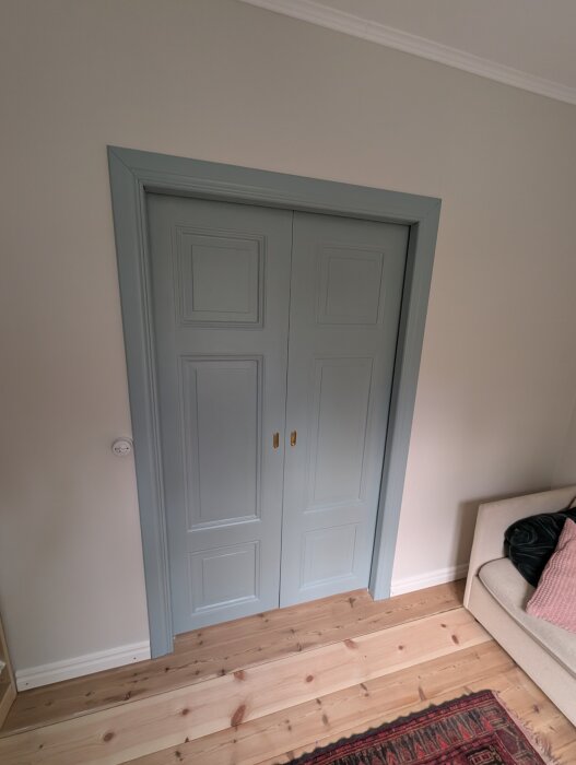 Dubbeldörr i ljusblått med guldfärgade handtag, del av en nyrenoverad matsal med trägolv och en del av en soffa synlig.