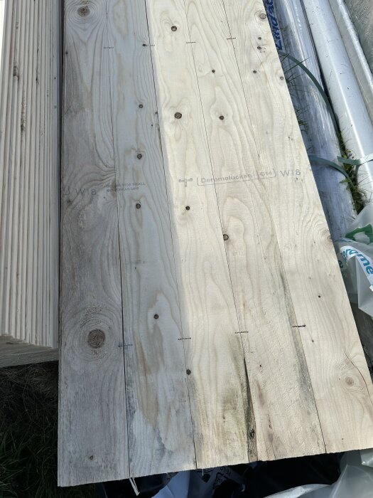 Torra och mögliga brädor staplade vid ett byggprojekt, med synlig trästruktur och märken, en blandning av fuktiga och torra ytor.