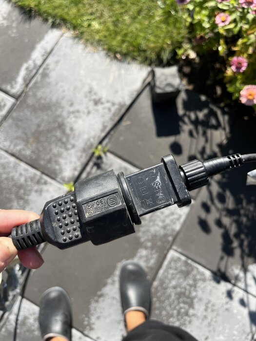 Närbild på en hand som håller en svart förlängningssladd med ett CE-märke, i en trädgård med plattor och blommor i bakgrunden.