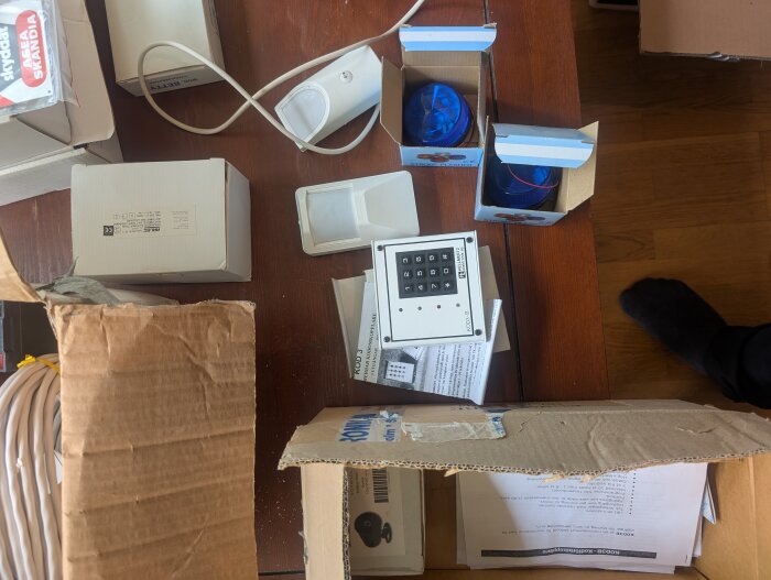 Lösa delar av ett gammalt larmsystem, inklusive en knappsats och sirener, är utspridda på ett bord bland kartonger och kablar.