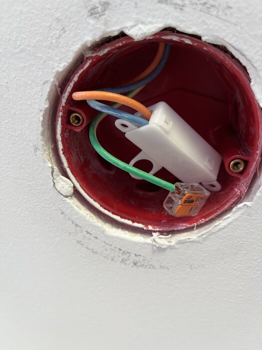 Rund infälld dosa i väggen med synliga elledningar i orange, grönt och blått, samt en WAGO-koppling, och infästningsgängor för lamputtag.