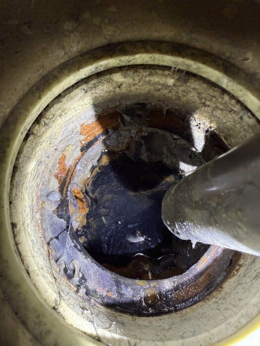 Rostig brun öppning på en brunn med ett rör som sträcker sig in. Brunnen är delvis täckt av smuts och fukt.
