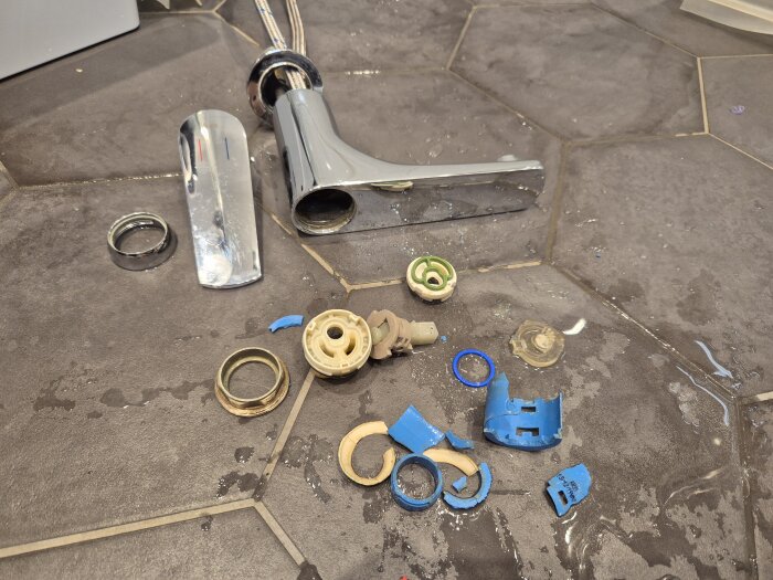 Demonterad tvättställsblandare med spruckna insatser och delar liggandes på ett vått, sjuhörnigt kakelgolv i badrummet.