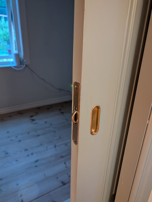 Närbild av dörr med nyinstallerade beslag i ett rum med trägolv och en sladd vid fönstret.