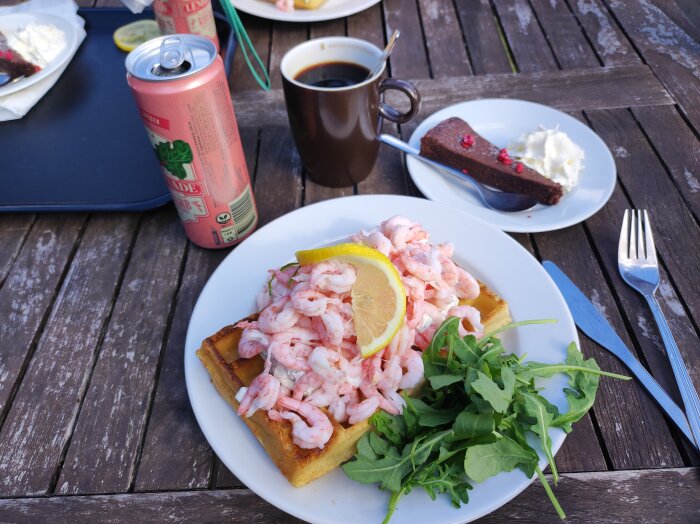 Belgisk våffla med skagenröra och räkor, en kopp kaffe, kladdkaka och vispgrädde på träbord utomhus.