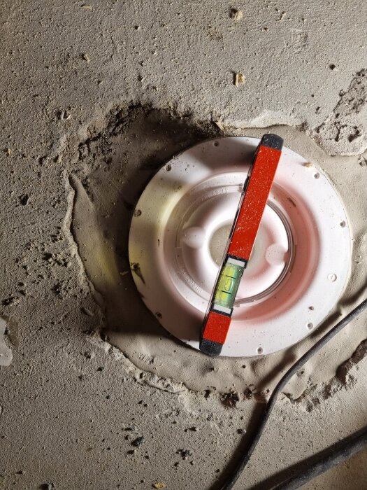 Rund ventil delvis installerad i golv med ett vattenpass placerat över, omgiven av byggdamm och smuts.