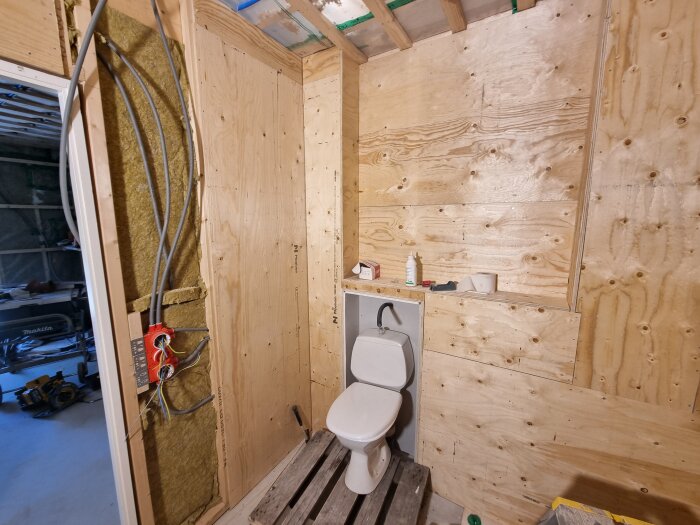 Byggplats med träskivor på väggarna, ett installerat toalettstol och exponerade rörledningar i ett halvfärdigt rum.