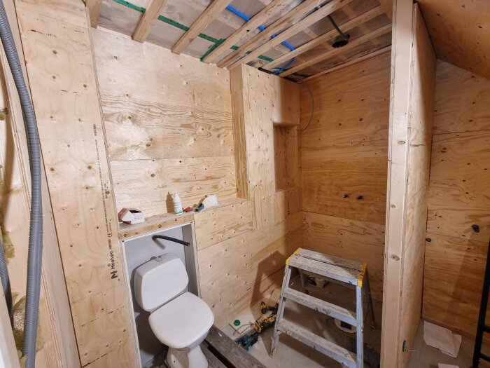 Oskyddat badrum med träpanel, toalett och stege, eluttag och verktyg i rummet. Förberett för elvärmegolvinstallation.