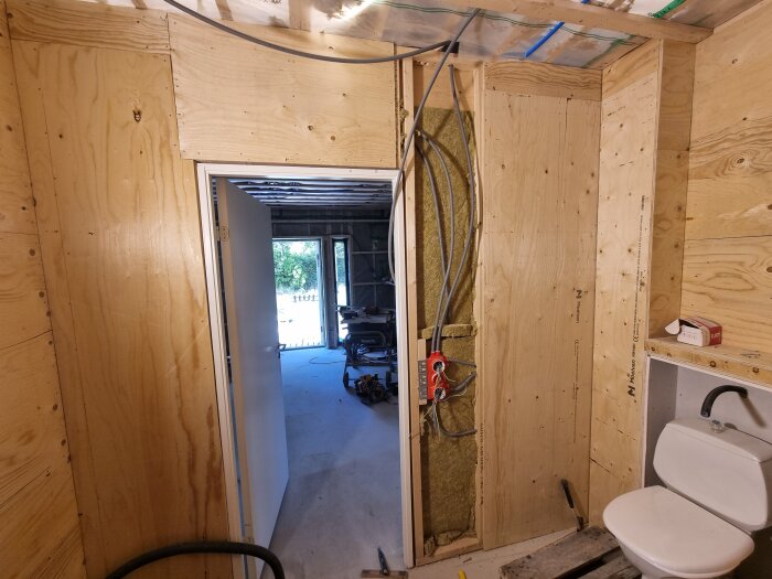 Byggarbetsplats med väggar klädda i träskivor, exponerade rör, och en toalett installerad. Dörr öppnar mot rum med utrustning.