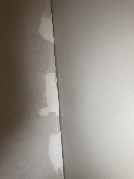 Närbild av ett vägg hörn som visar ojämnt spacklat område med gips och ett gap mellan angränsande väggar utan skarvremsa.