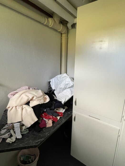 En hög med kläder på en mörk köksbänk i ett hörn med vitmålade rör på väggen och ett vitt skåp bredvid.