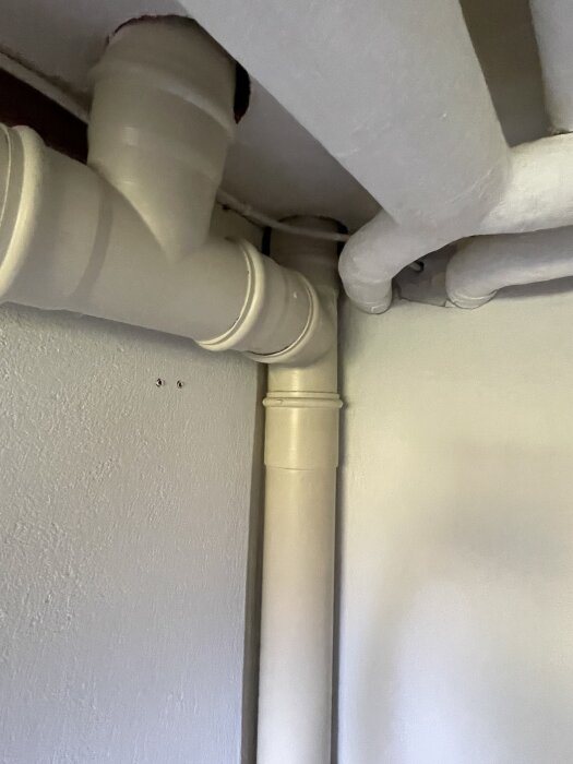 Rörsystem vid ett takhjärn i en byggnad, där flera vita rör, inklusive ett T-förgreningsrör, går längs väggen och det vita taket.