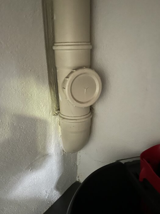 Vit avloppsledning med inspektionslucka monterad mot en vit vägg, bredvid en delvis synlig svart och röd plastbehållare.