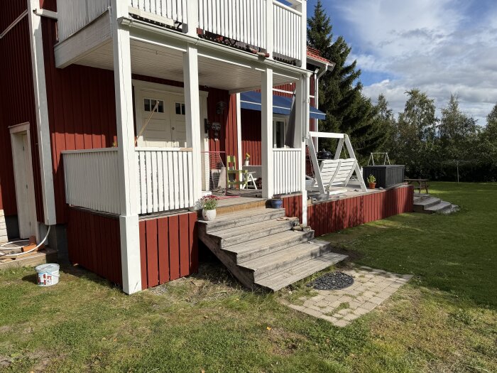 Röd verandatrappa nymålad i solsken, med vita staket och omgivande grönska. Målarburk vid sidan av trappan.