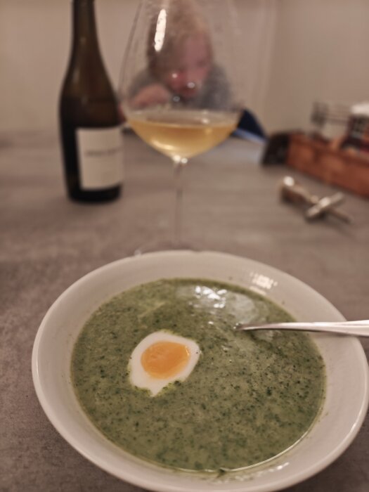 Grön spenatsoppa med ett halvt kokt ägg i en skål, ett glas vin och en suddig person i bakgrunden.