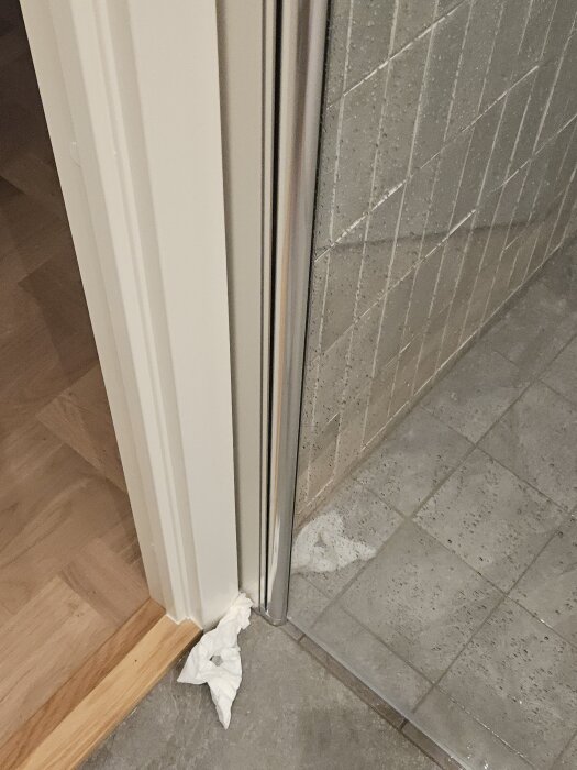 Duschdörr med vattenläckage vid golv; fuktigt golv och papper placerat för att suga upp vatten. Tröskel syns i bakgrunden.
