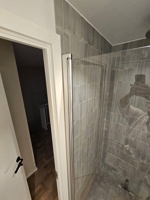 Duschhörna med glasvägg, grå kakel och en öppen dörr mot hallen. Reflektion av en person synlig i glaset.