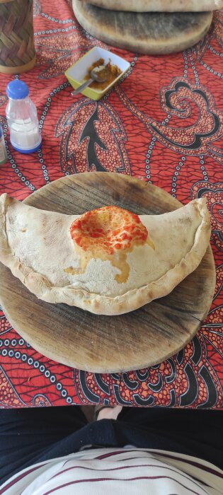 En nygräddad calzone-pizza på träunderlägg, med ett rött, mönstrat bordstyg och några tillbehör i bakgrunden.