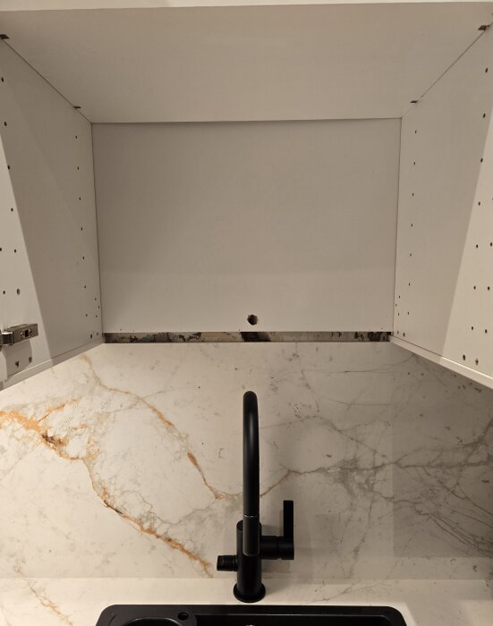 Öppet skåp ovanför diskho med rund hål i hyllan, och marmorliknande stänkskydd bakom svart kran.